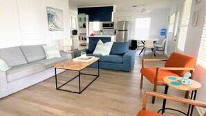 Un salon moderne avec un canapé gris, un canapé bleu et des fauteuils orange. On aperçoit un coin cuisine et une table basse en bois à l’arrière-plan.