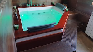 Una vasca idromassaggio rettangolare con base in legno e piano in vetro, circondata da una parete rivestita di piastrelle scure.