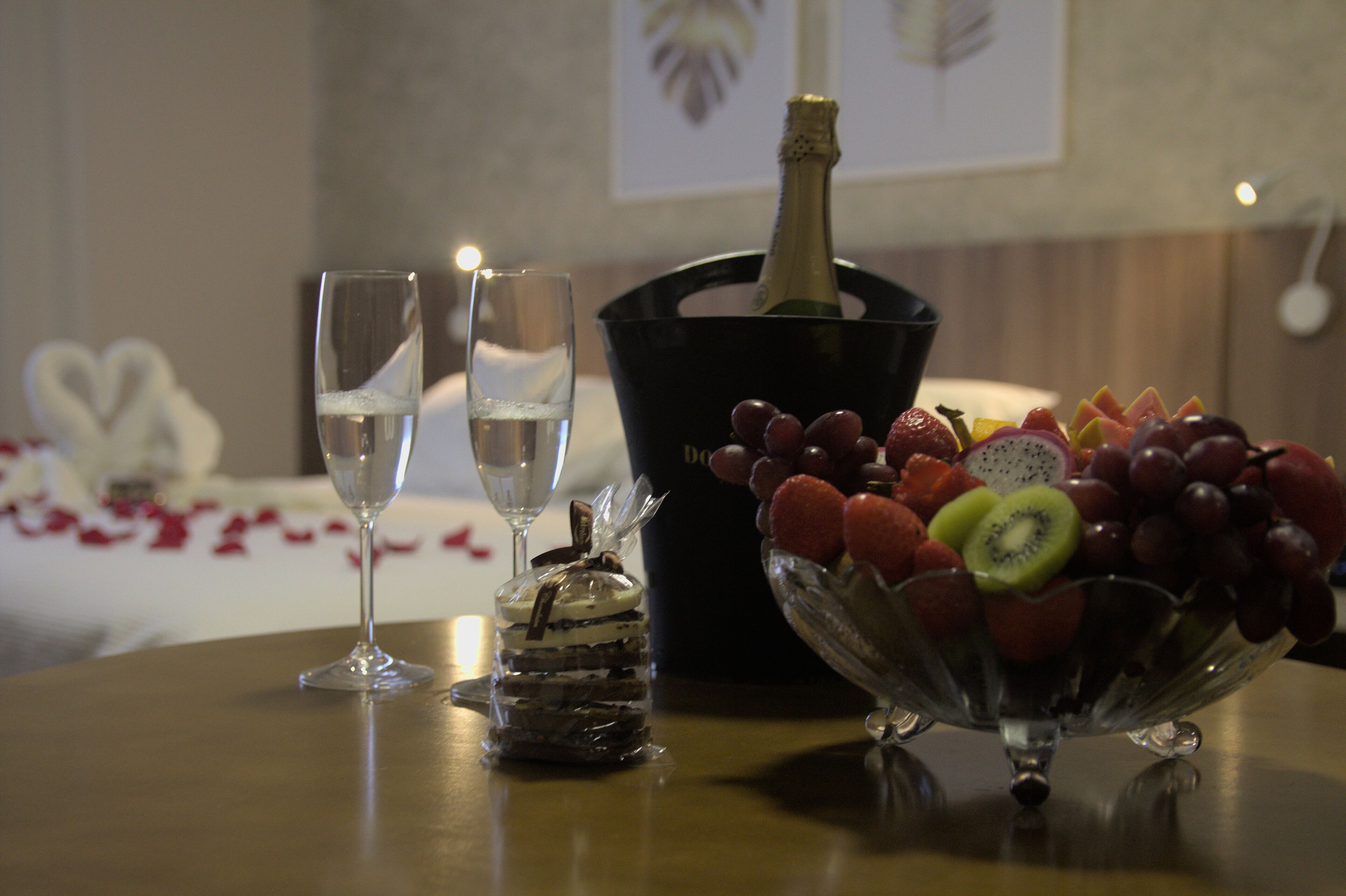 A table with two champagne flutes, a bottle of champagne, a fruit bowl, and a dessert in a glass jar.