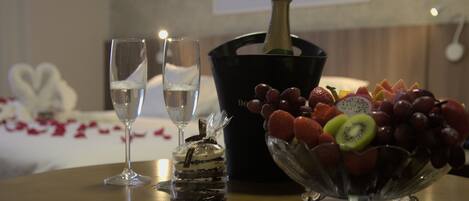 A table with two champagne flutes, a bottle of champagne, a fruit bowl, and a dessert in a glass jar.