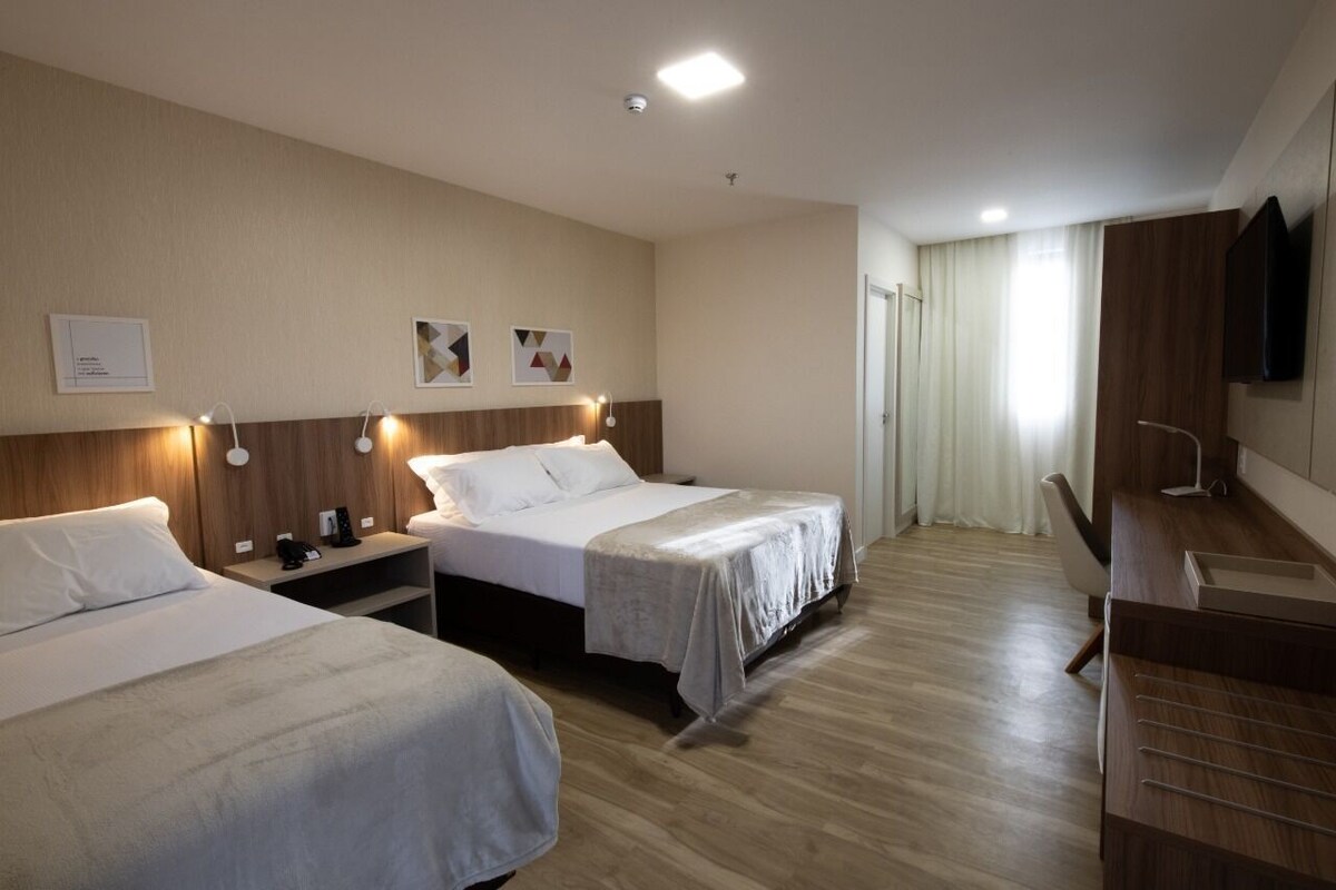 A hotel room with two beds, a wooden headboard, bedside lamps, a flat-screen TV, and a bathroom with a shower and toilet.