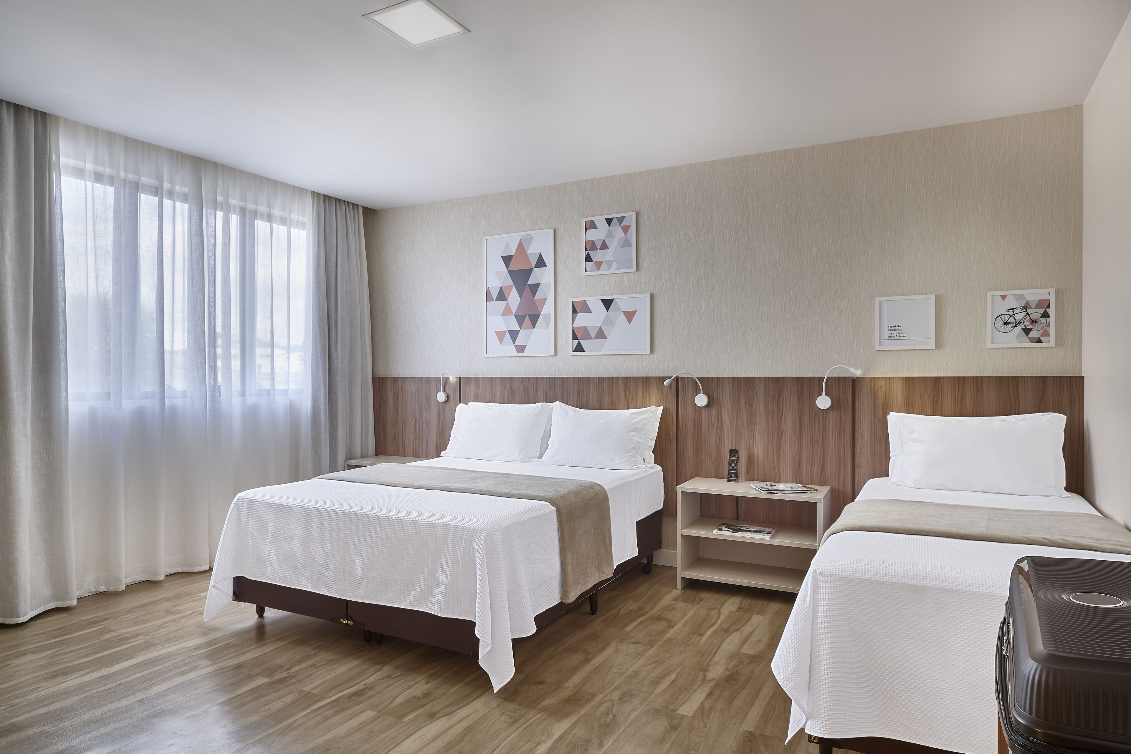 Quarto de hotel com duas camas, piso de madeira, uma janela grande com cortinas leves e três obras de arte penduradas na parede.