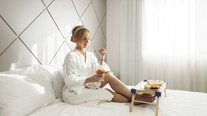 Une personne vêtue d’une robe blanche est assise sur un lit, une table d’appoint contenant de la nourriture et une boisson se trouve à proximité.