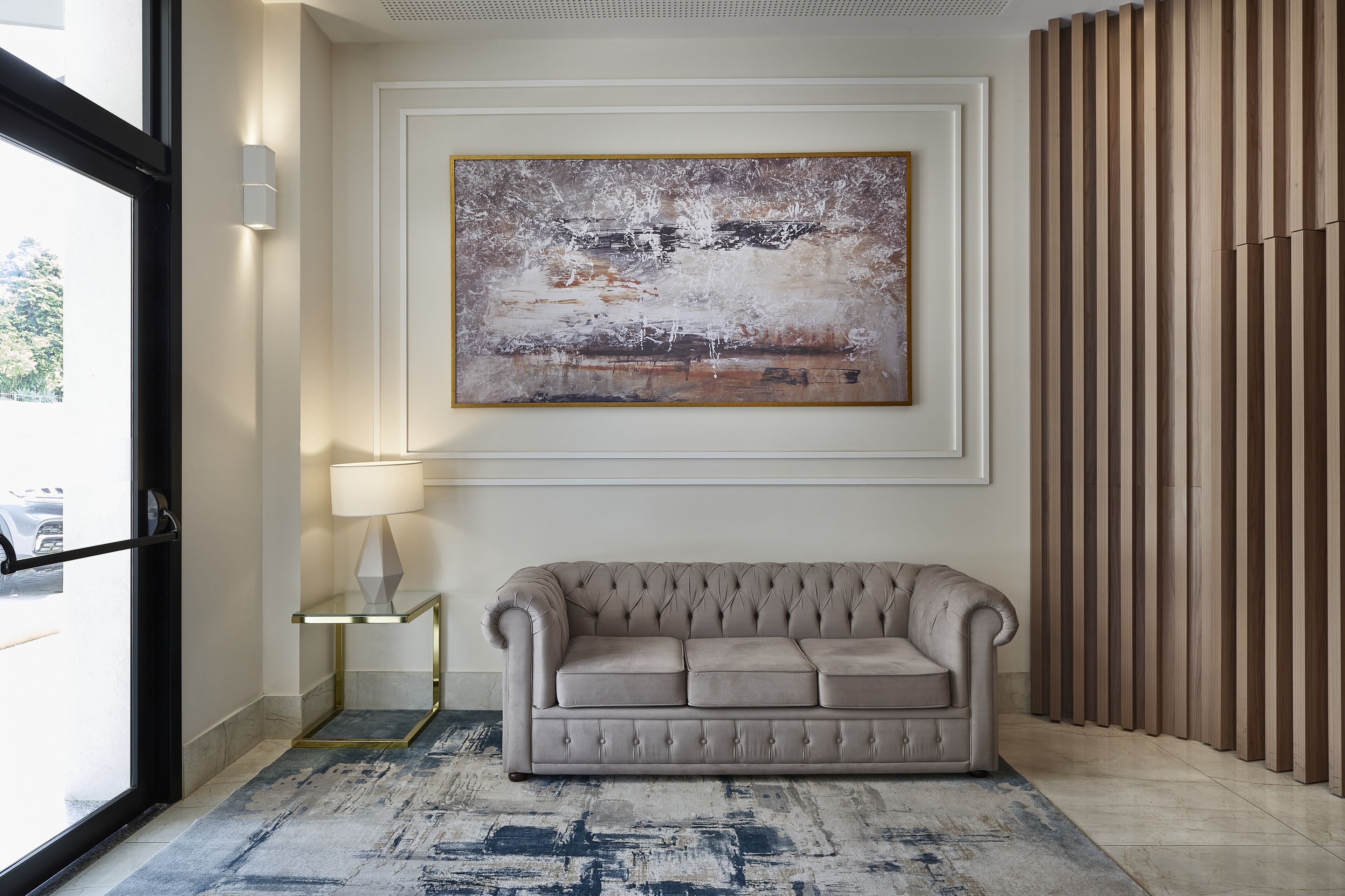 Una hall moderna con un divano color beige, un quadro astratto di grandi dimensioni, una lampada e un pannello di legno a rivestimento.