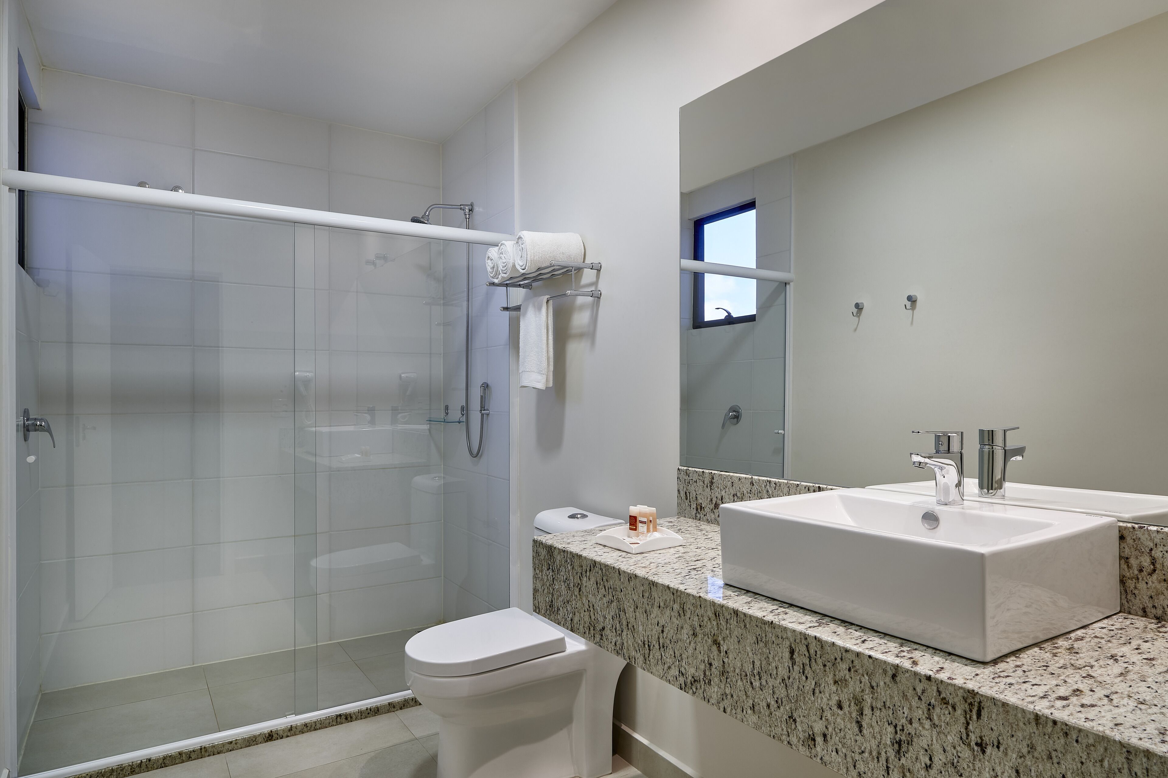 Un baño moderno con mampara de vidrio, inodoro blanco, lavamanos grande con encimera de granito y un espejo sobre el lavamanos.