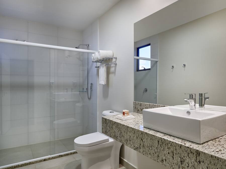 Un baño moderno con mampara de vidrio, inodoro blanco, lavamanos grande con encimera de granito y un espejo sobre el lavamanos.