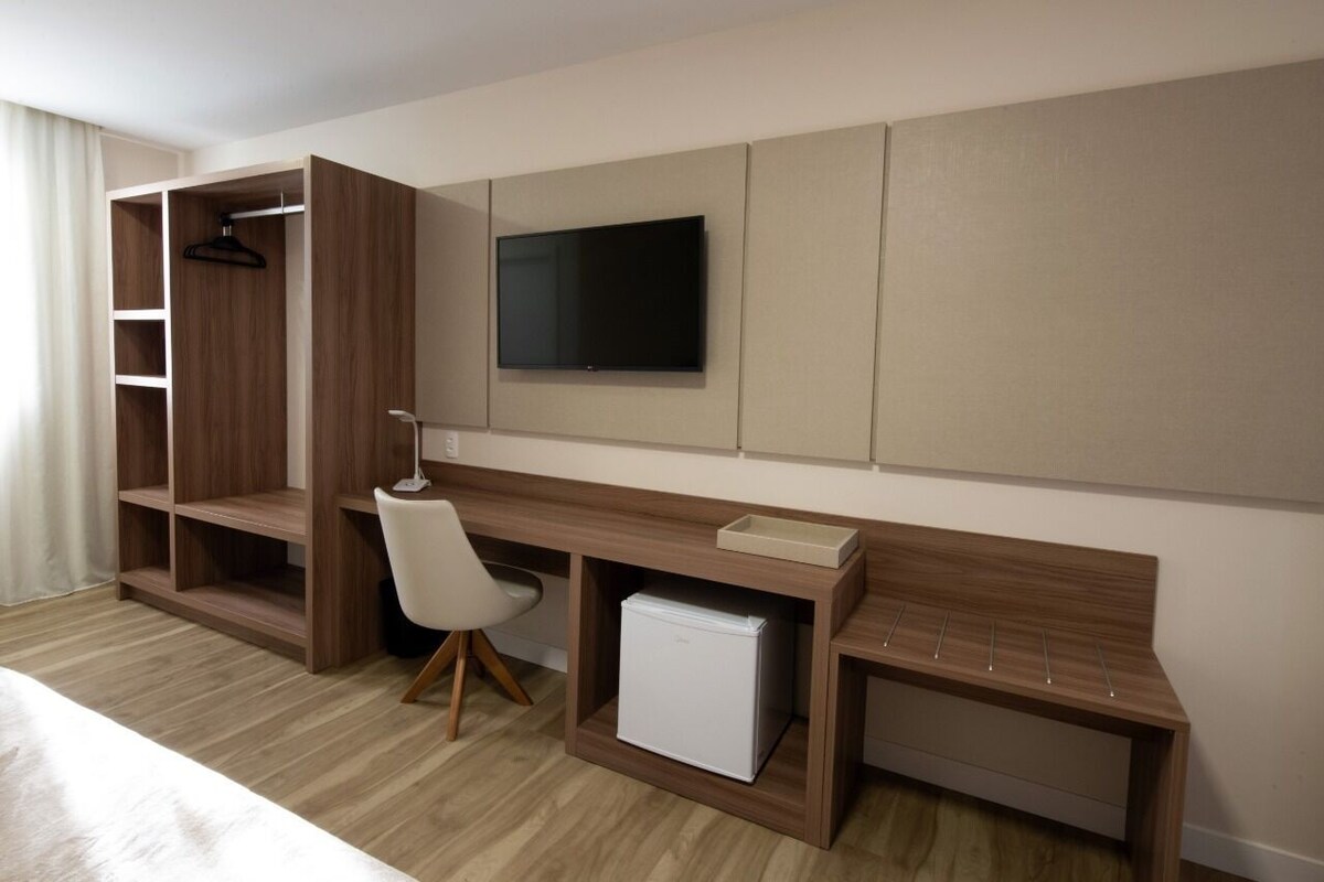 A hotel room with a bed, a desk with a chair, a TV, a wardrobe, and a mini-fridge.