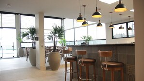 Un espace bar moderne, doté de nombreux tabourets et d’un éclairage suspendu. On y trouve de grandes plantes en pot et un grand vase sur le comptoir.