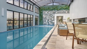 Une piscine intérieure moderne dotée d'un toit en verre, offrant une vue sur la ville, et d'un espace détente avec des coussins.