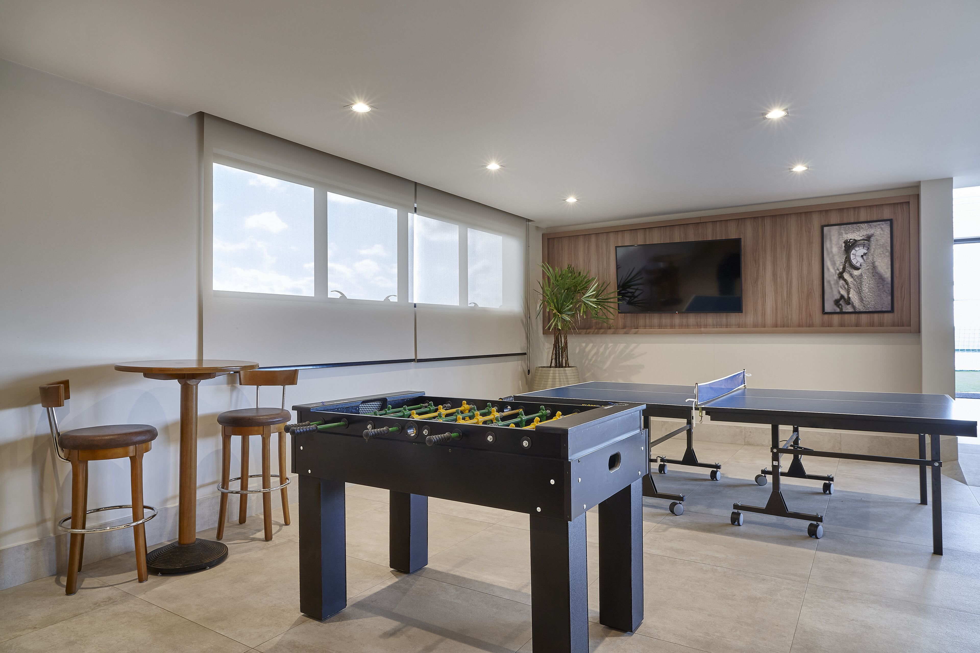 A room with a foosball table, two bar stools, a ping pong table, and a large window.