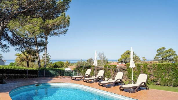 Ein Pool mit Liegestühlen und Sonnenschirmen, umgeben von Grünpflanzen und Blick aufs Meer.