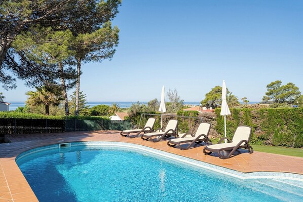 A swimming pool with lounge chairs and umbrellas, surrounded by greenery and a view of the sea.