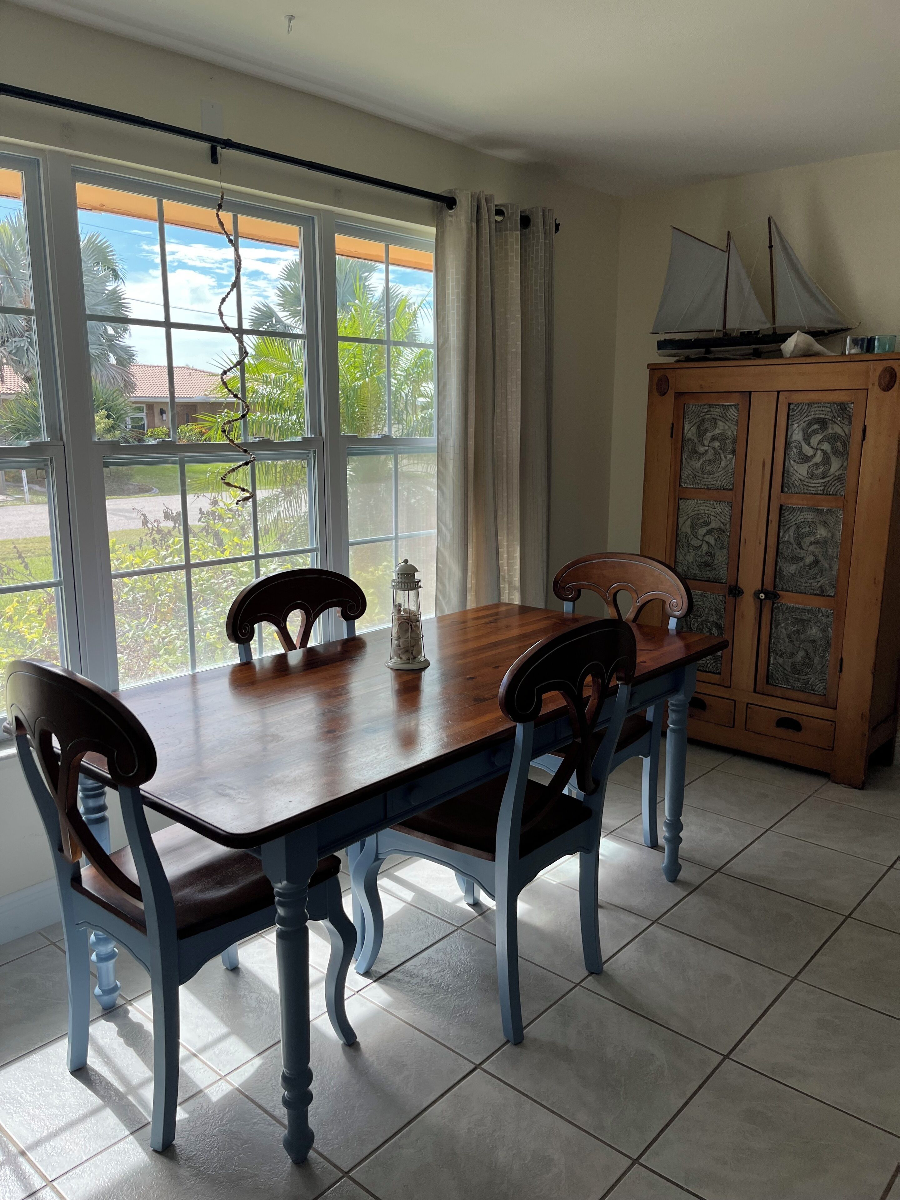 A dining area with a wooden table and chairs, a cabinet with decorative panels, and a view of the outdoors through large windows.