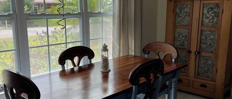 A dining area with a wooden table and chairs, a cabinet with decorative panels, and a view of the outdoors through large windows.