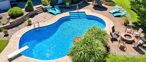 Una piscina con tobogán integrado, rodeada por un patio de ladrillo, sillas de descanso y una zona con chimenea.