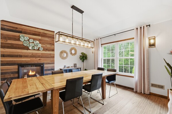A dining area with a wooden table, black chairs, a fireplace, and a large window with curtains. - Incredible House in the Heart of the Poconos. Private Heated Pool & Hot Tub (Long Pond)