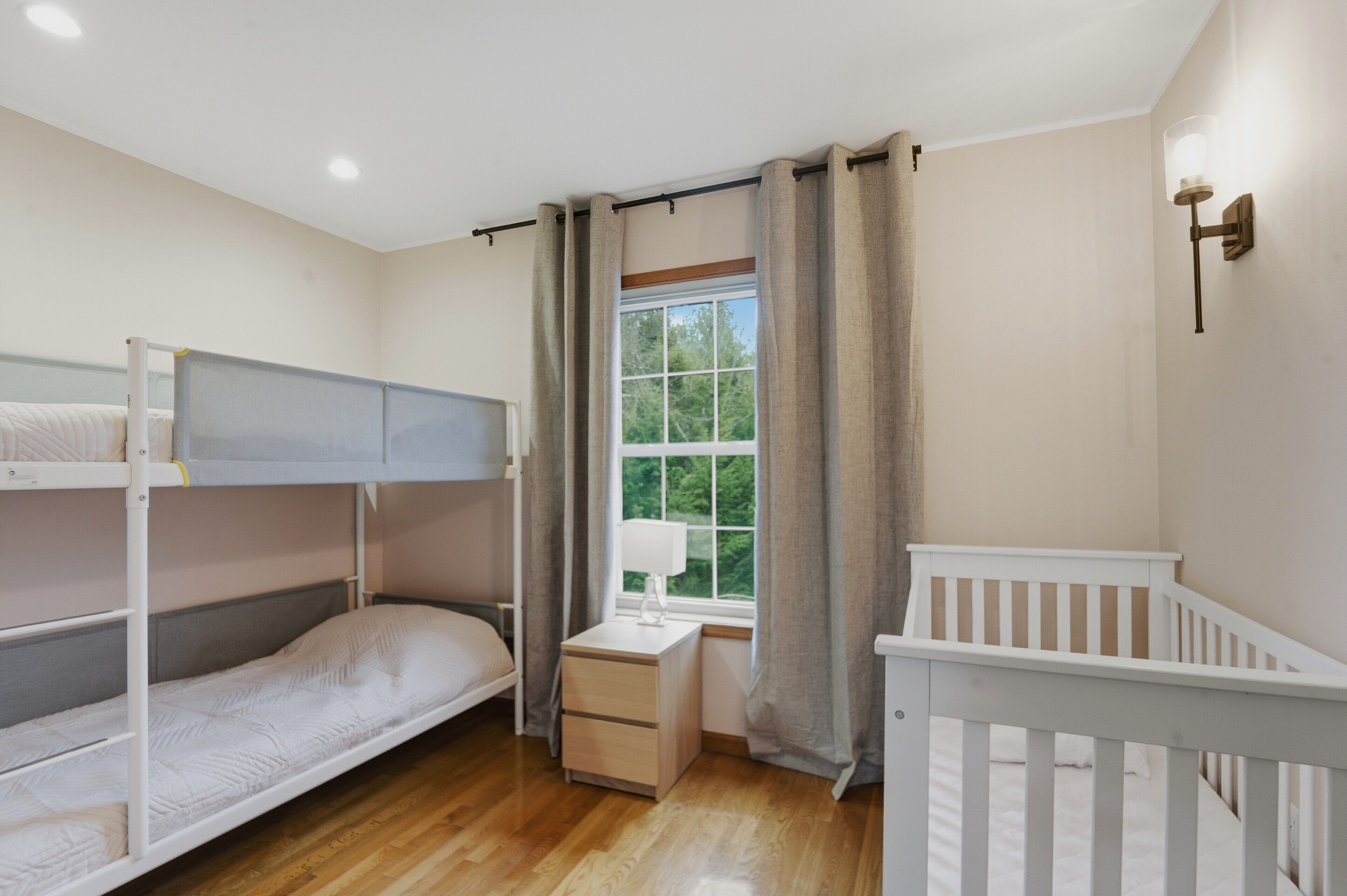 A room with bunk beds, a bedside table with a lamp, a window with curtains, and a white crib.
