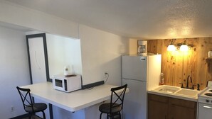 A compact kitchen with a white countertop, two bar stools, a microwave, a refrigerator, a sink with a wooden backsplash, and a stove.
