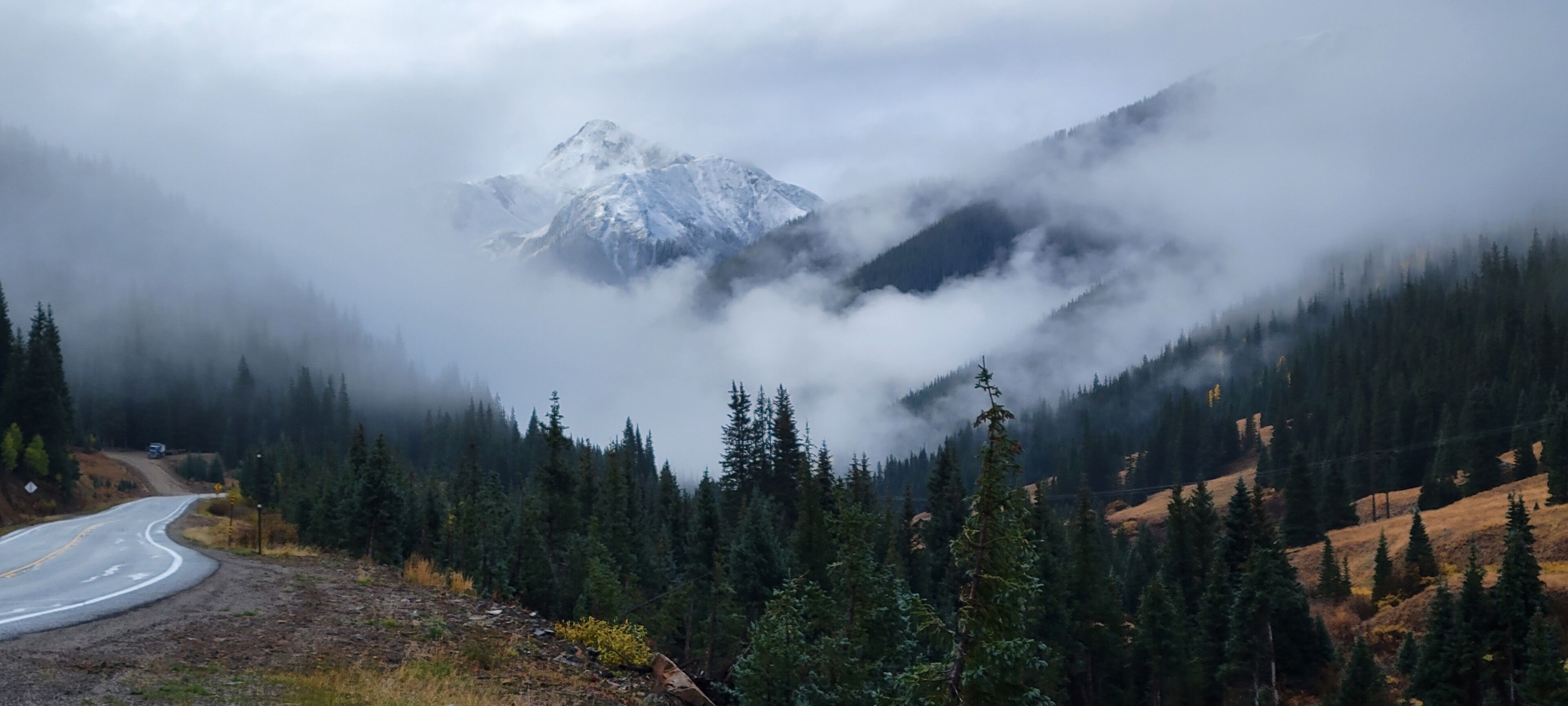 A winding road through a forested mountain area with a snow-capped peak in the background.