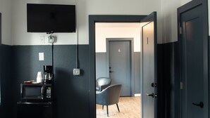 A hotel room with a flat-screen TV mounted on the wall, a coffee maker, a microwave, and a chair. The room has a door with the number 6 on it, leading to another room.