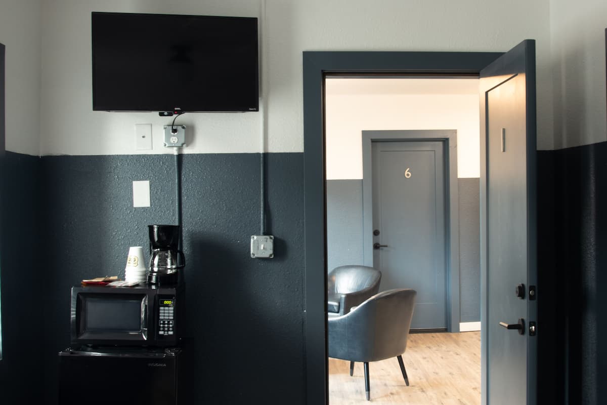 A hotel room with a flat-screen TV mounted on the wall, a coffee maker, a microwave, and a chair. The room has a door with the number 6 on it, leading to another room.