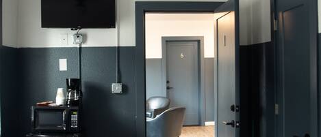 Ein Hotelzimmer mit Flachbildfernseher an der Wand, einer Kaffeemaschine, einer Mikrowelle und einem Sessel. Das Zimmer hat eine Tür mit der Nummer 6, die in einen weiteren Raum führt.