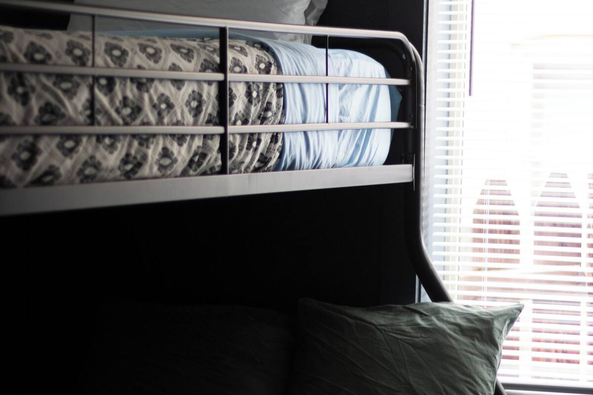 A bunk bed with patterned bedding and pillows, a window with blinds, and a plain wall.