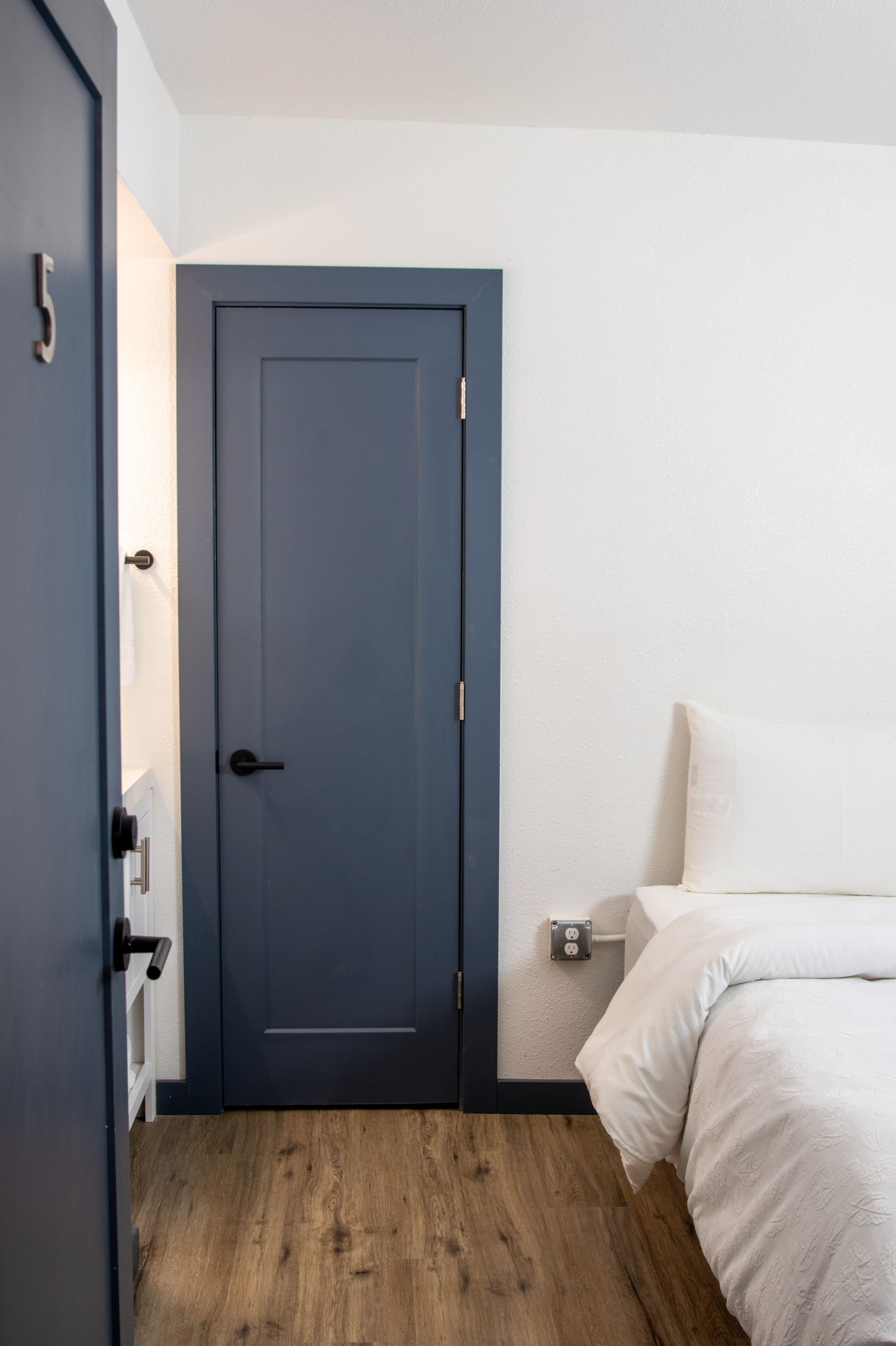 A room with a dark blue door, a bed with white bedding, and a wooden floor.