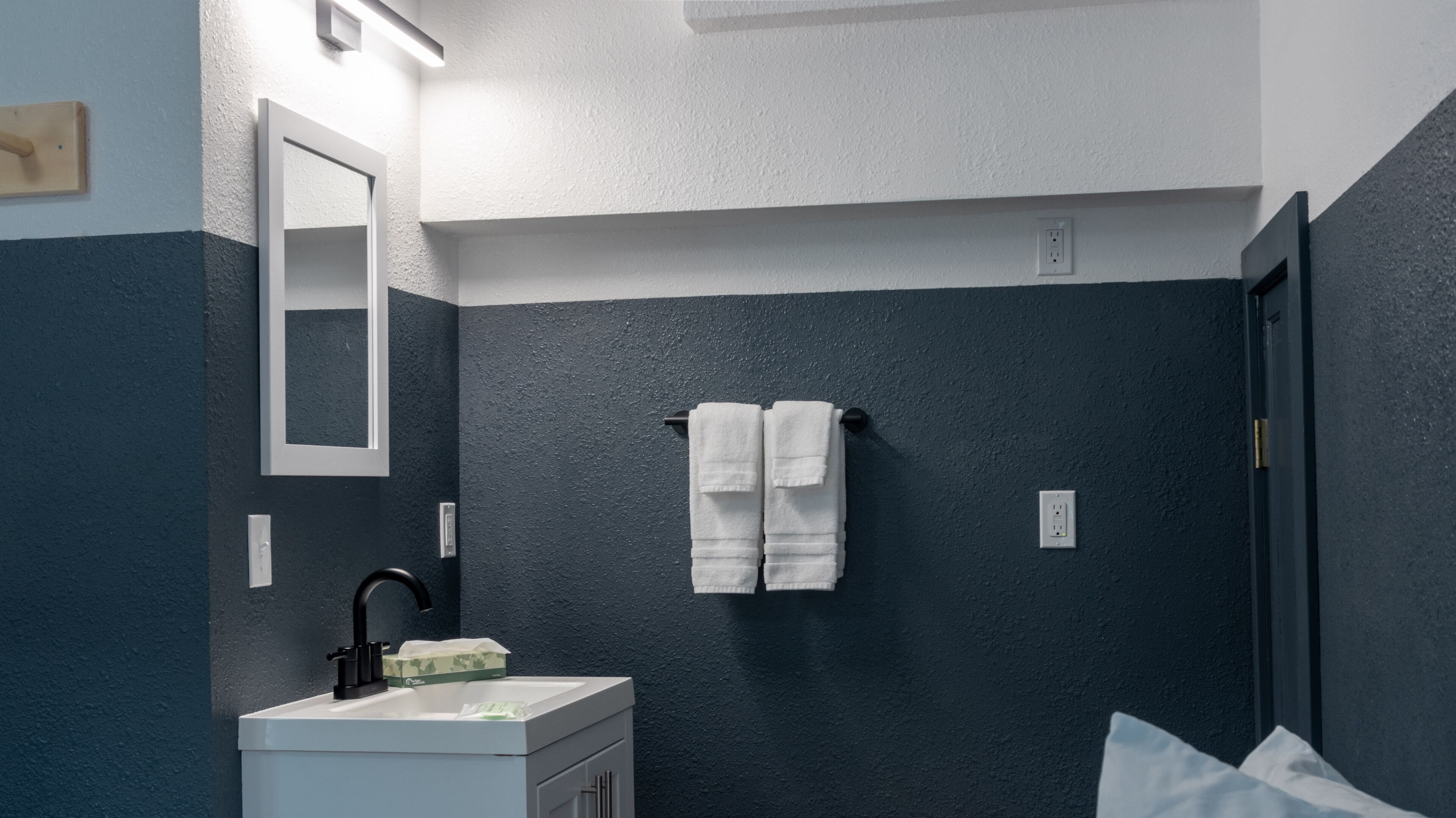 Une salle de bain avec un mur bleu foncé, un lavabo blanc, un robinet noir, un miroir et un porte-serviettes avec deux serviettes blanches.