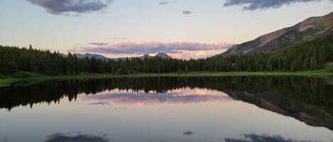 A calm lake reflecting trees and mountains, surrounded by a forested area.
