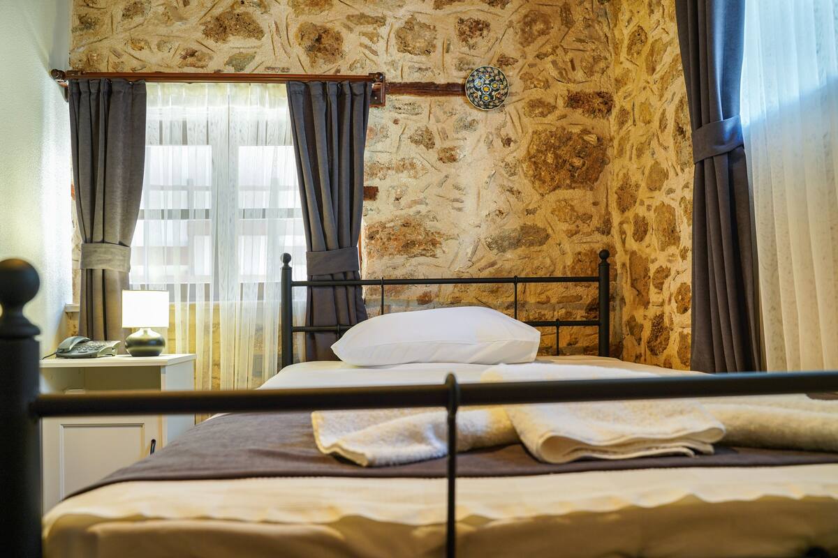 A bed with a headboard, a bedside table with a lamp, a telephone, and a wall clock.