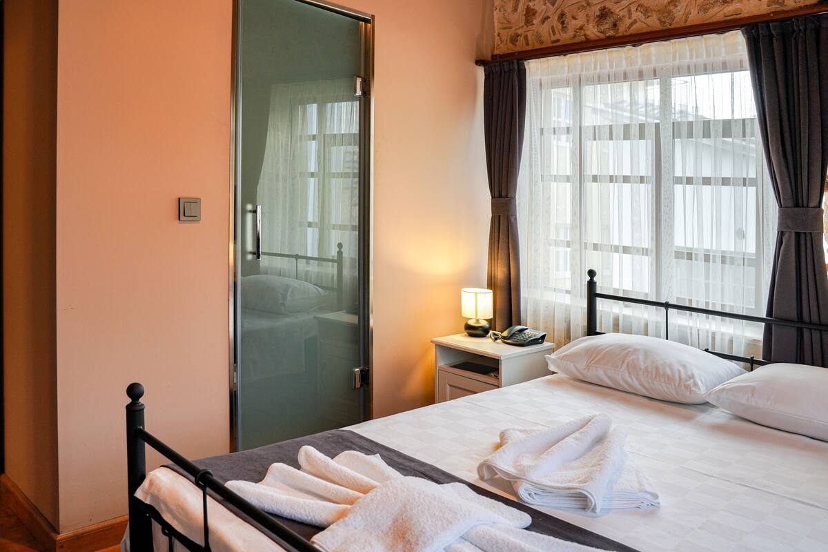 A hotel room with a bed, a nightstand with a lamp, a telephone, and a door with a glass panel.