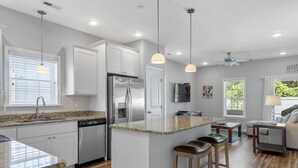 A modern kitchen with white cabinetry, stainless steel appliances, and a granite countertop. There are bar stools and a dining area with a wooden table and chairs. - The E Gull Has Landed: Private Spa, Shared Pool (Kill Devil Hills)