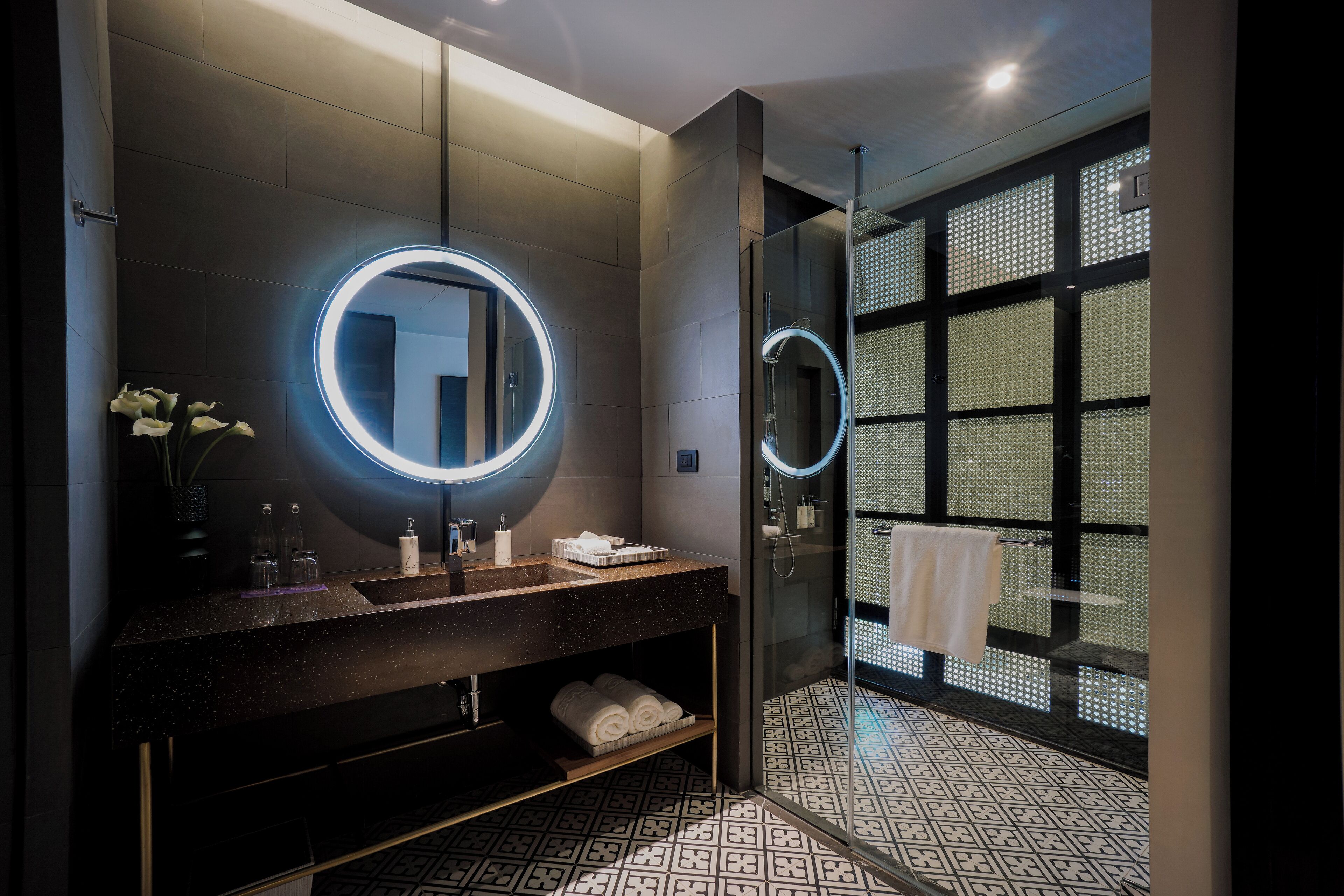 A modern bathroom with a dark granite countertop, a large mirror with a circular light, a glass shower enclosure, and patterned floor tiles.