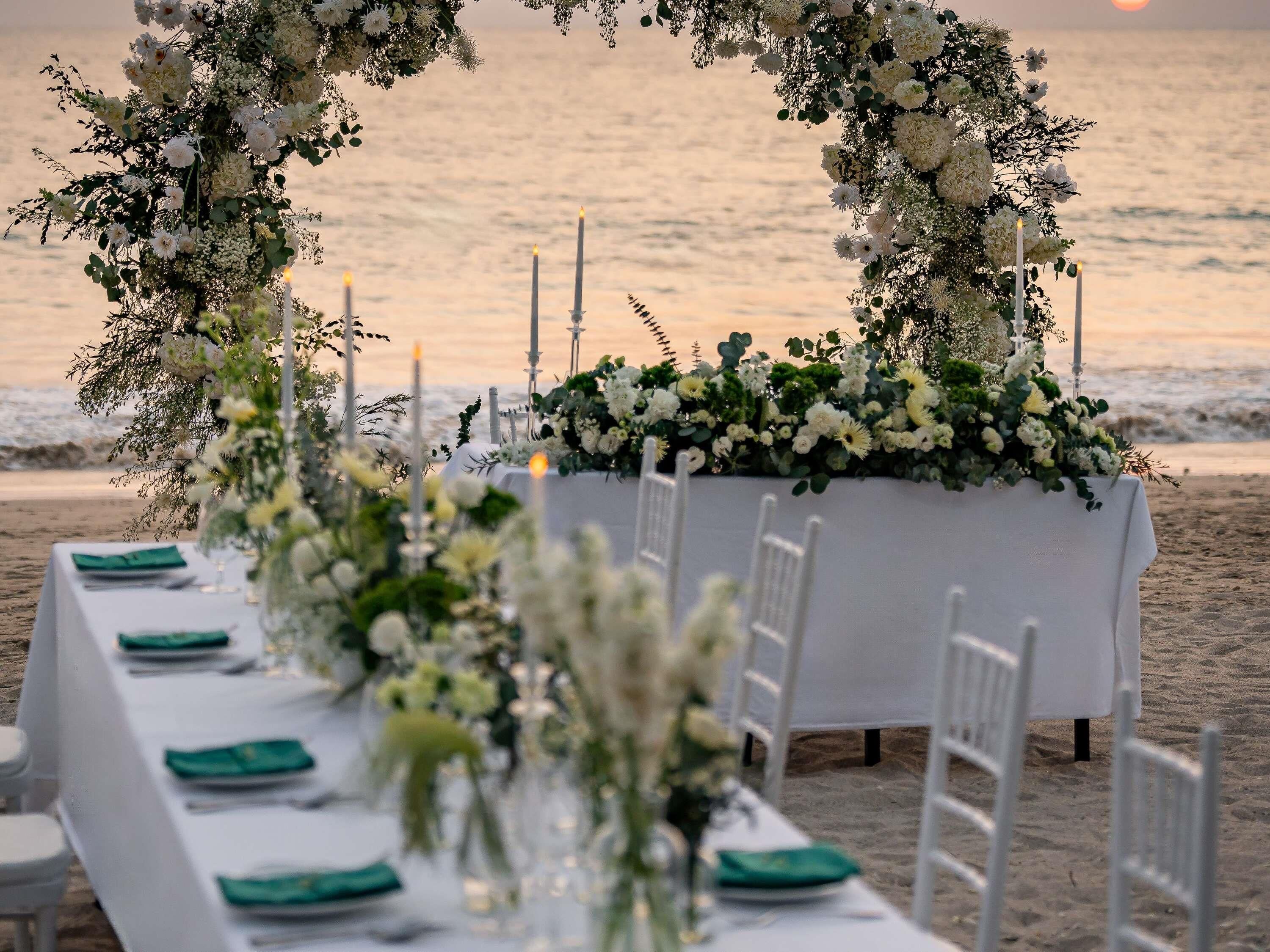 A beachside table setup with white tablecloths, green napkins, and floral arrangements.