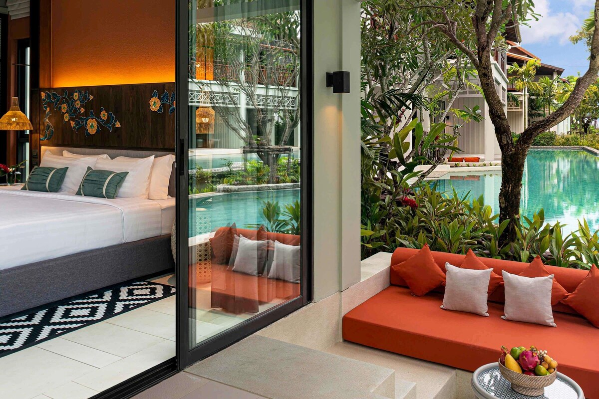 A modern hotel room with a bed, a glass wall, a pool, and a seating area with cushions.
