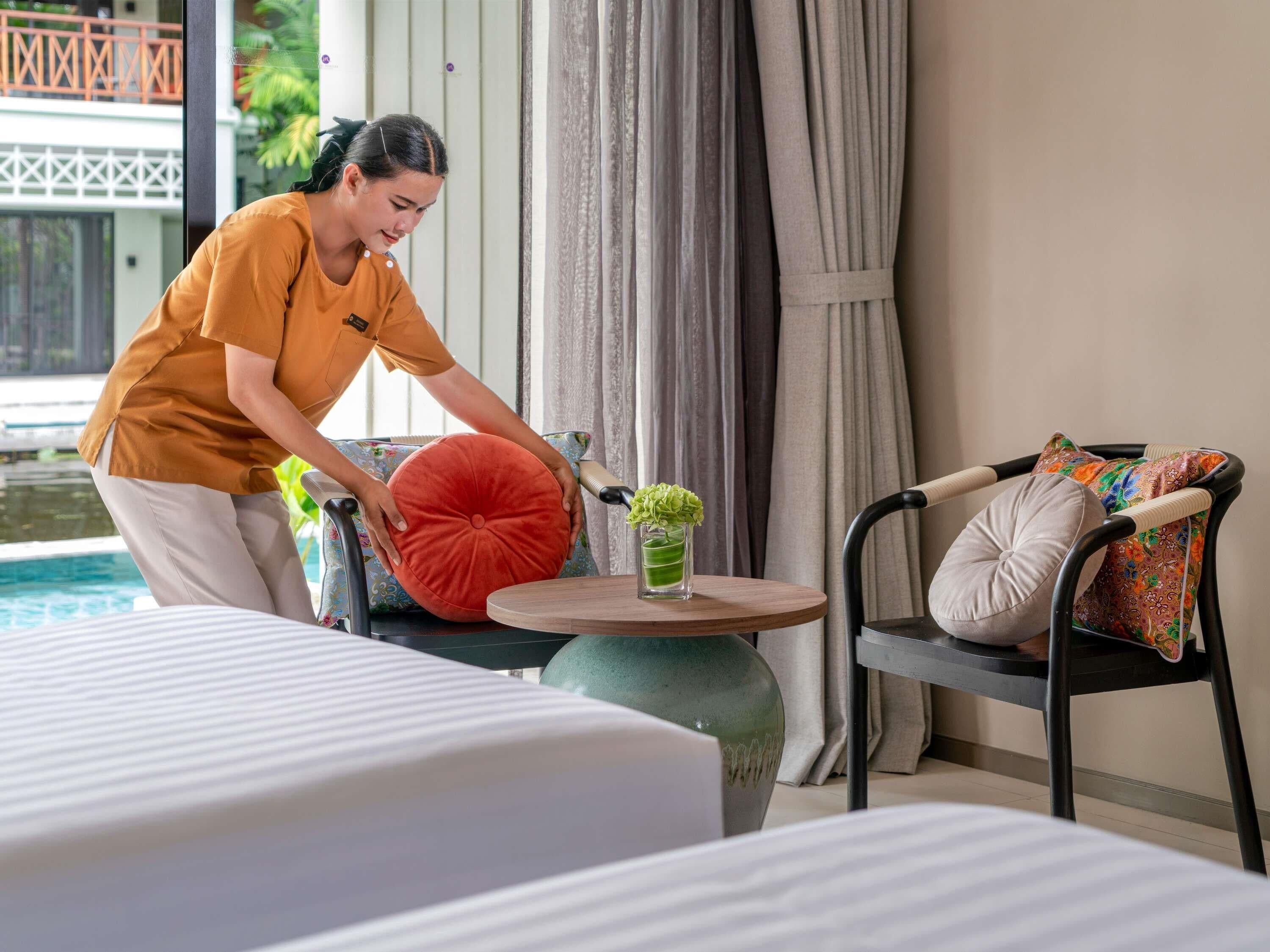 Habitación de hotel con una cama, una mesita redonda con un florero, una silla con cojín y vista a una piscina.