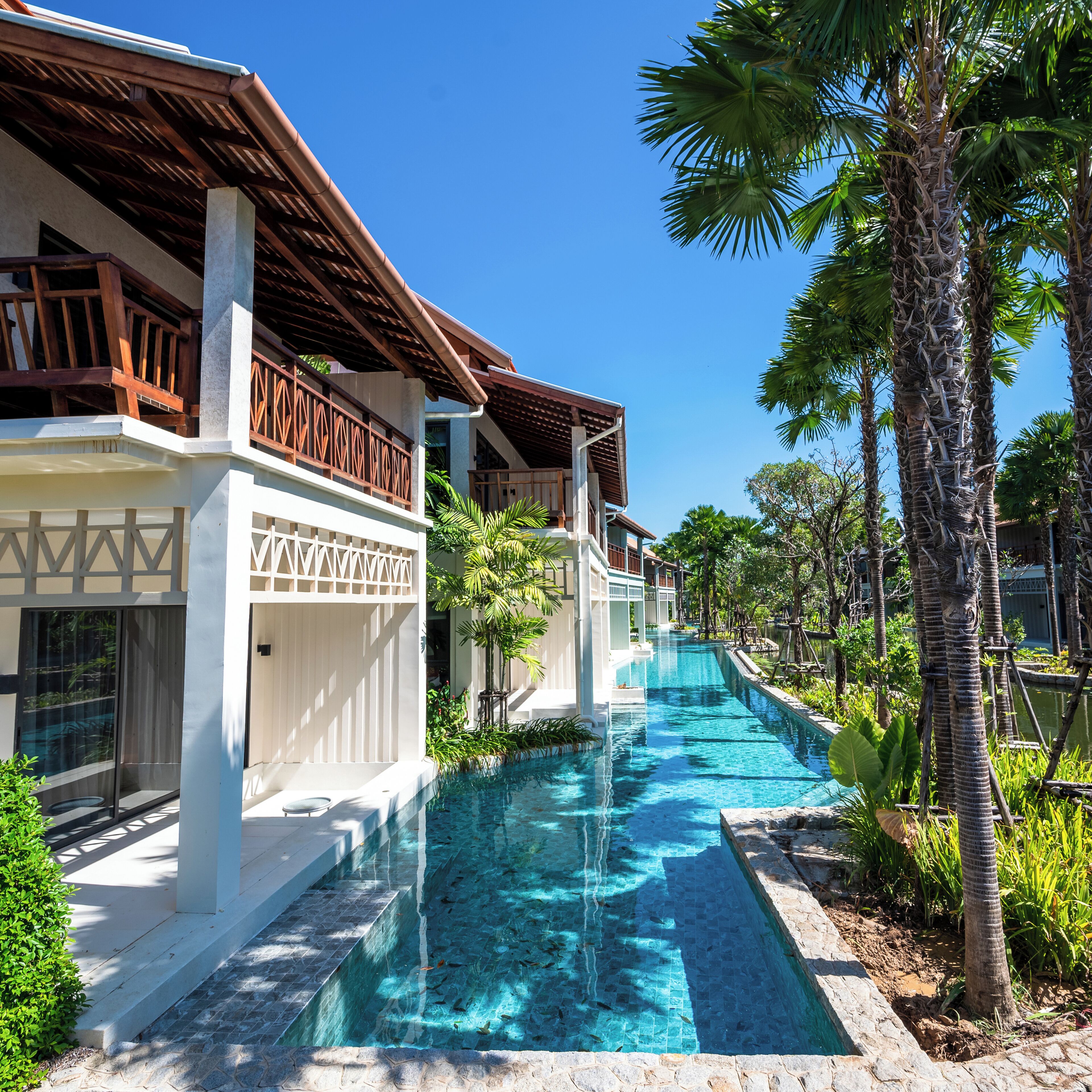 Um resort com piscina, palmeiras e um edifício moderno com varanda de madeira.