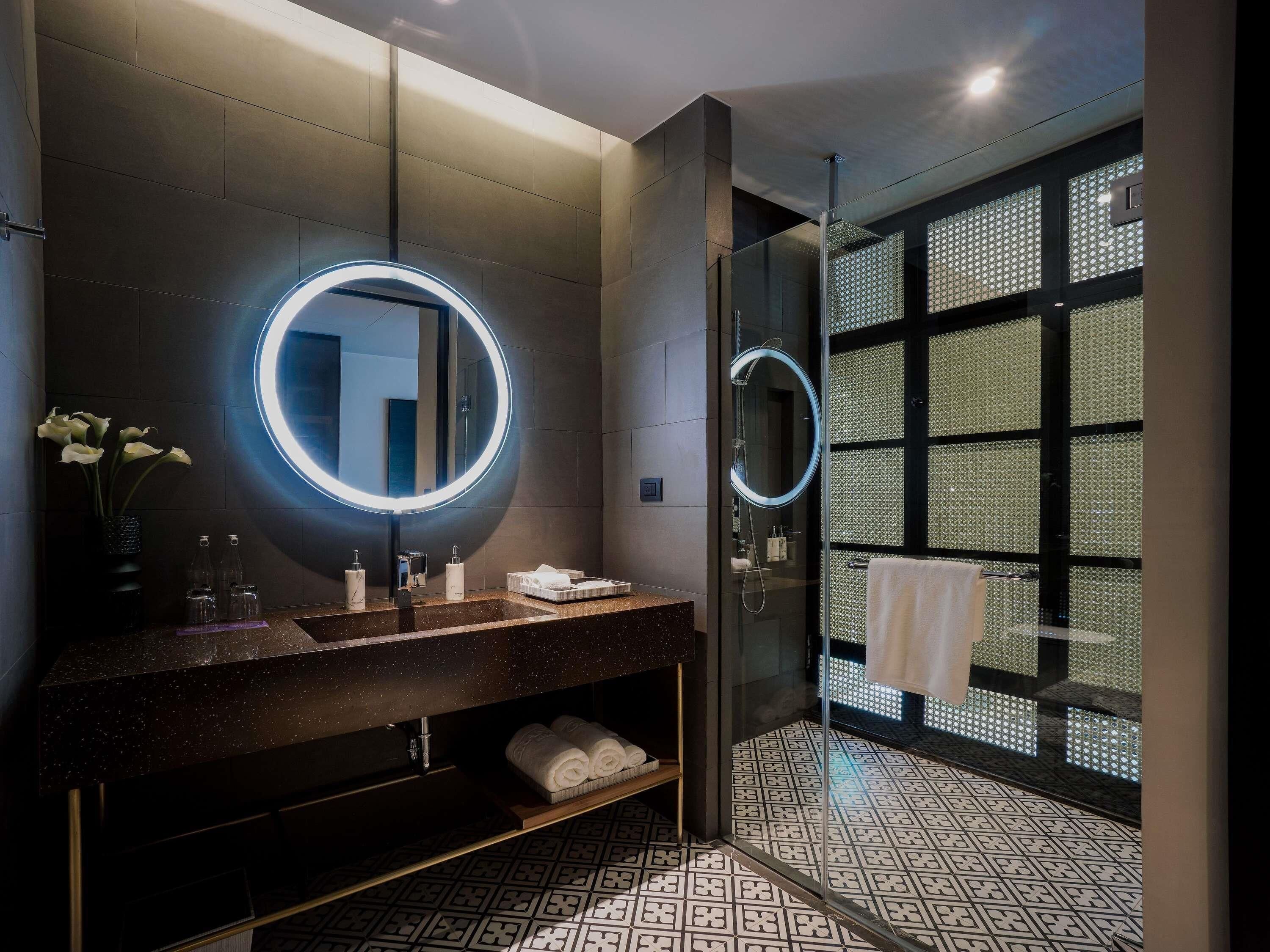 A modern bathroom with a round mirror, a glass shower enclosure, and patterned floor tiles.