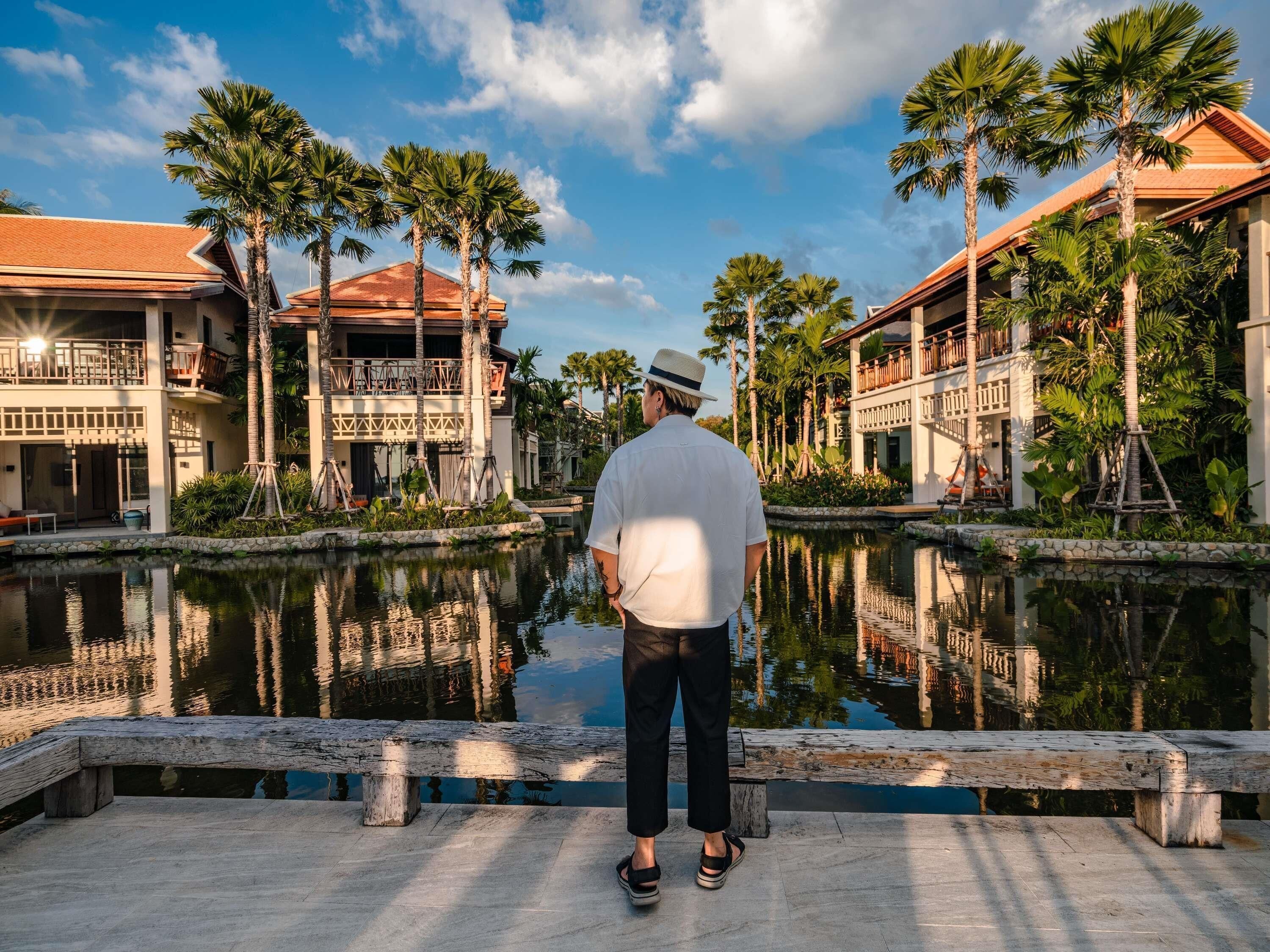 A man standing by a reflective water body with buildings and palm trees in the background.