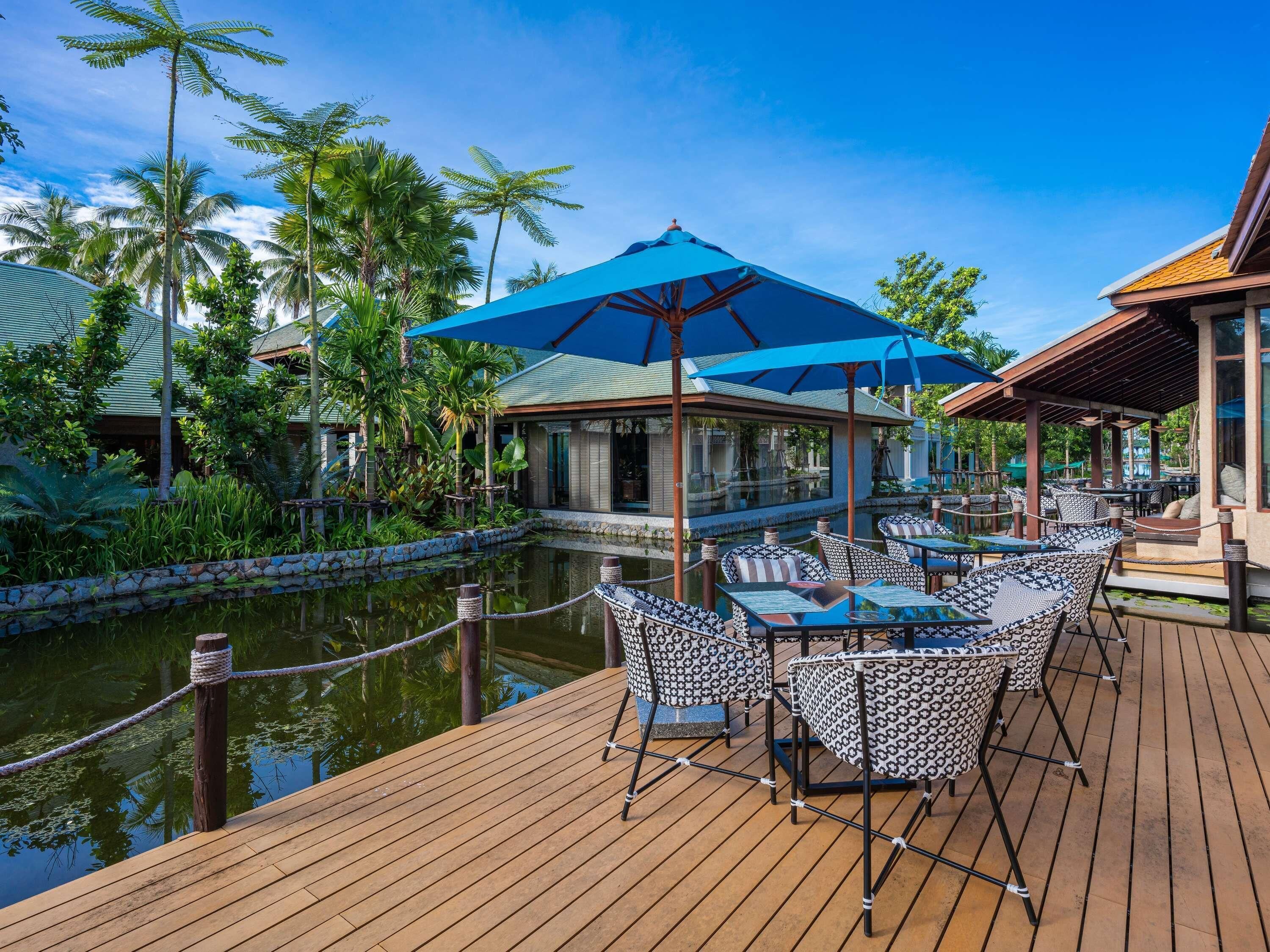 A wooden deck with tables and chairs, a pool, and a building with a blue roof.