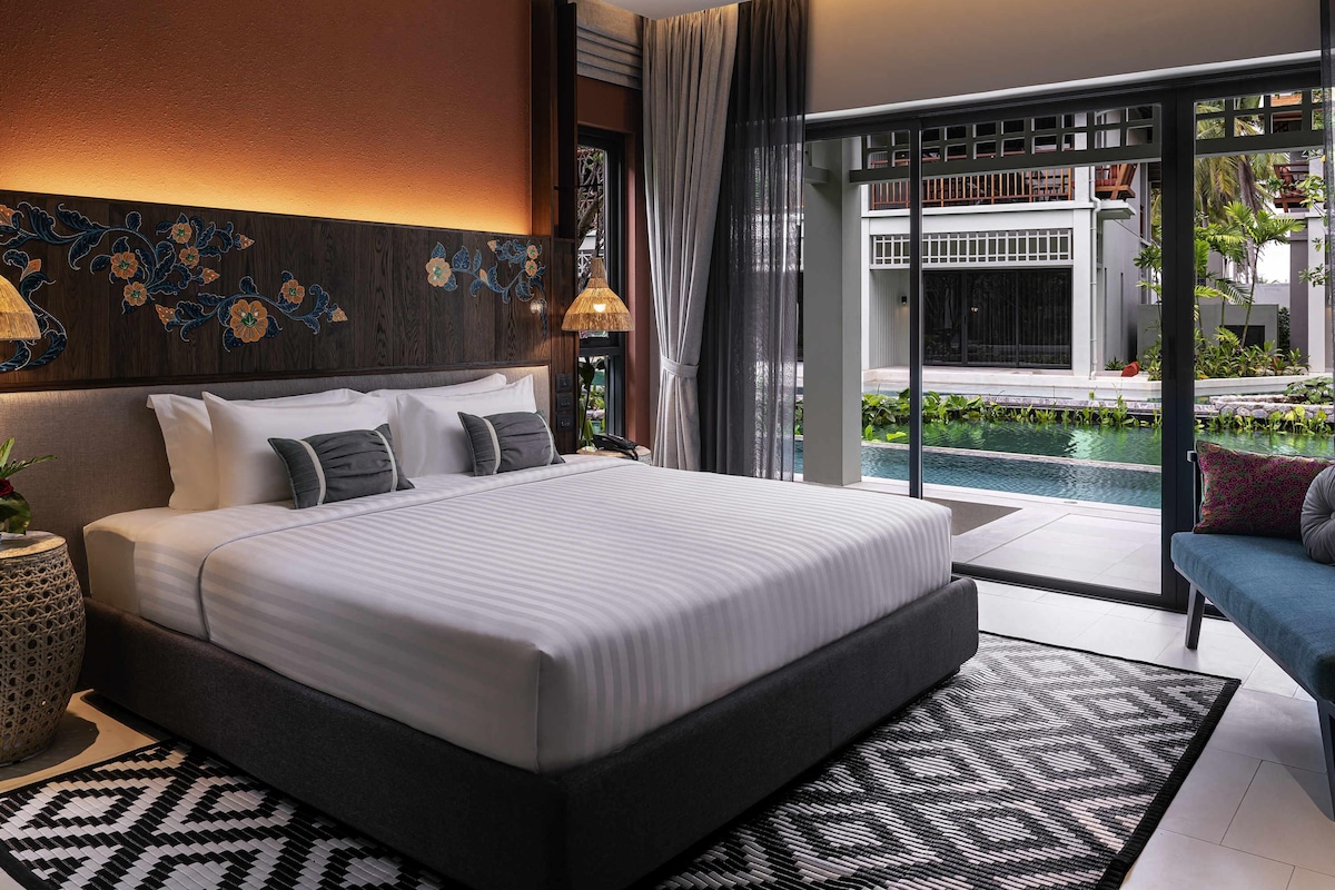 Una habitación de hotel moderna con una cama grande, vista a una piscina y un balcón con mobiliario exterior.