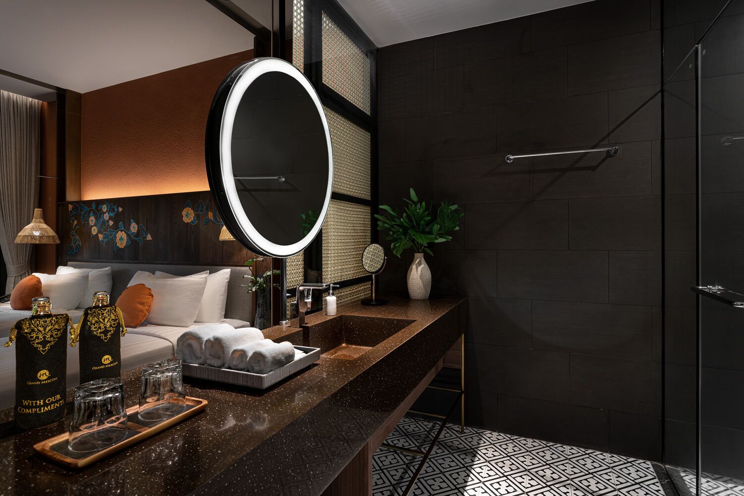 A modern bathroom with a dark tiled wall, a round mirror, a sink with a tray of towels, and a shower area.