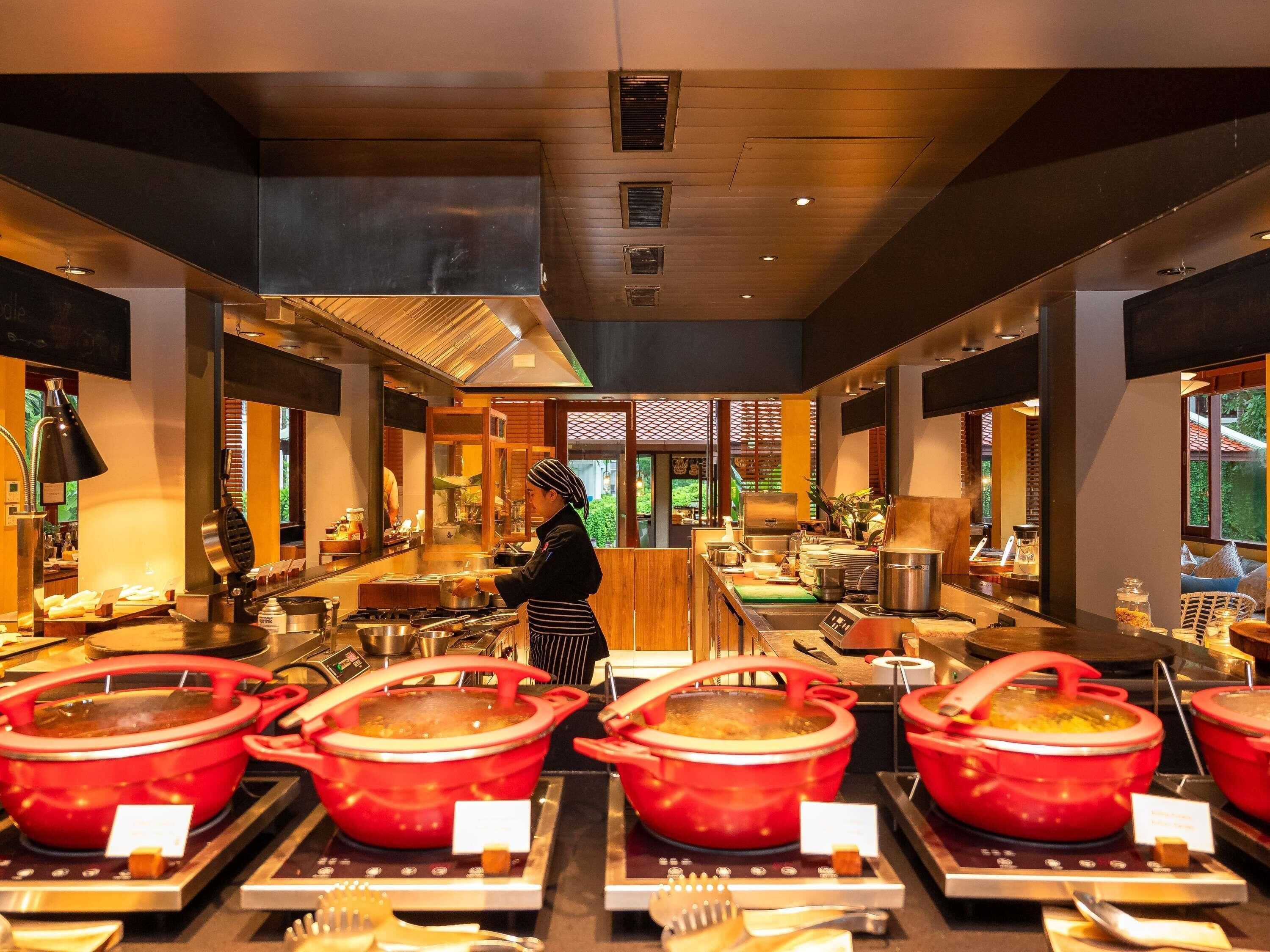 Un restaurante con varias ollas rojas en la barra, un cocinero preparando comida y vistas al exterior a través de amplias ventanas.
