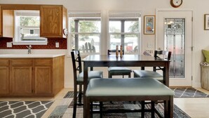 A kitchen with wooden cabinets, a dining table with four chairs, and a bench. There is a window with blinds, a door with a glass panel, and a clock on the wall. - River Hideaway Retreat (Southport)
