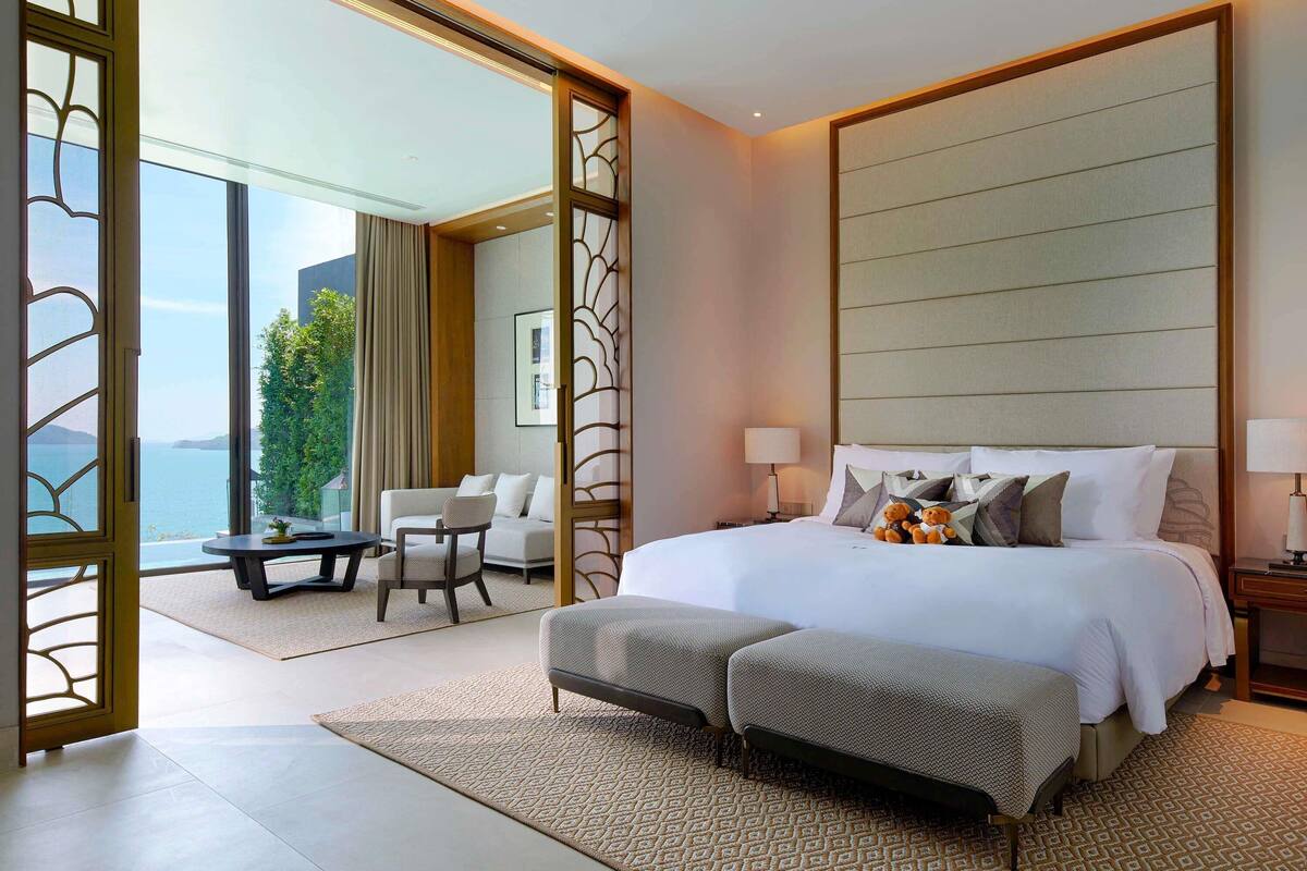 A modern hotel room with a large bed, a sofa, a dining table, and a balcony with a view of the sea.