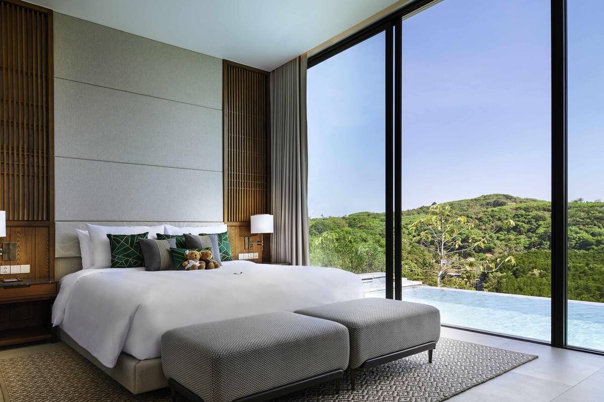 A modern hotel room with a large bed, a bench, a view of a lush landscape, and a pool.