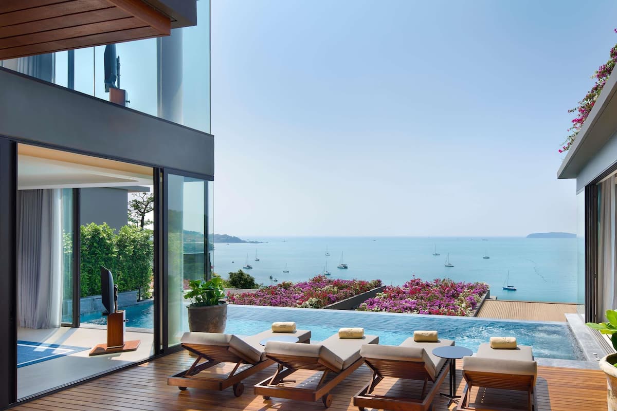 A modern outdoor lounge area with a pool, wooden deck, and a view of the sea with boats.
