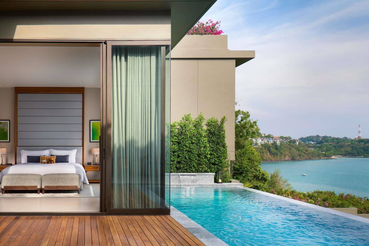 A modern hotel room with a large bed, a glass door leading to a balcony with a pool overlooking a scenic coastline.