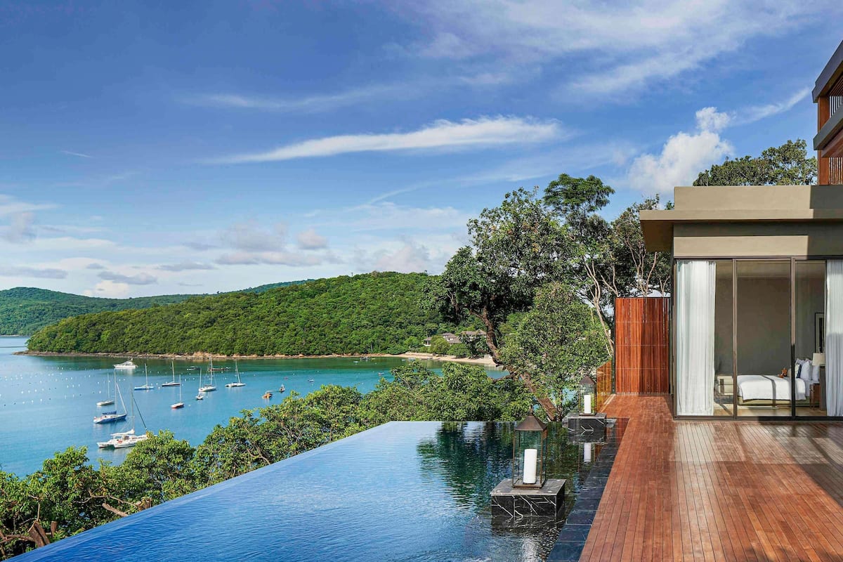A modern infinity pool overlooking a bay with boats, a wooden deck, and a hillside.